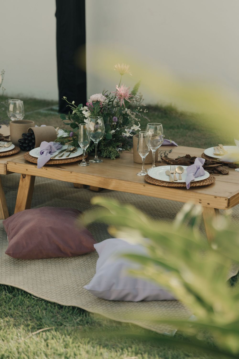 Elegant outdoor picnic setup with plush pillows, wooden serving boards, and delicate table settings on grass.