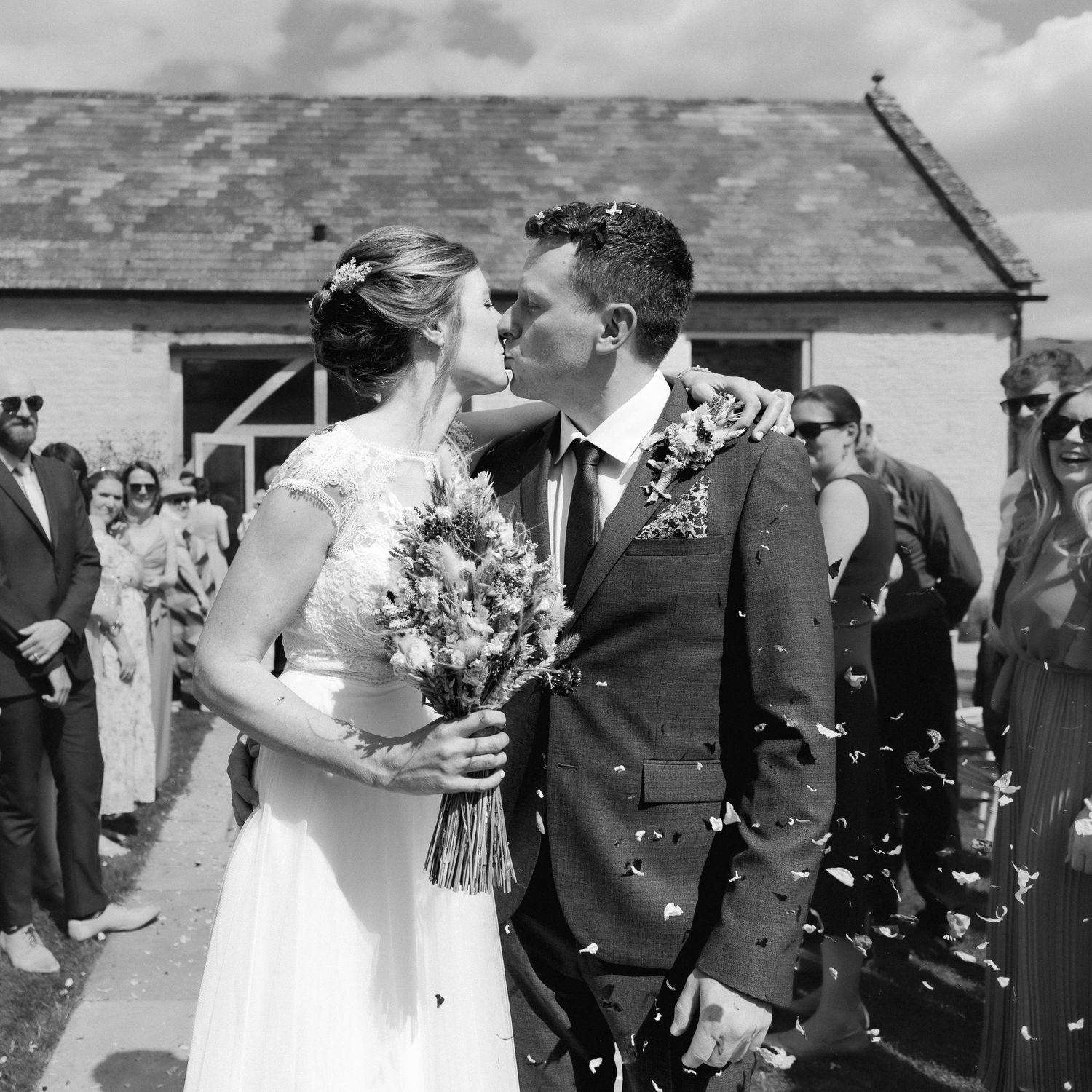 A black and white wedding photograph showing a romantic moment between newlyweds amid falling confetti outside a rustic building.