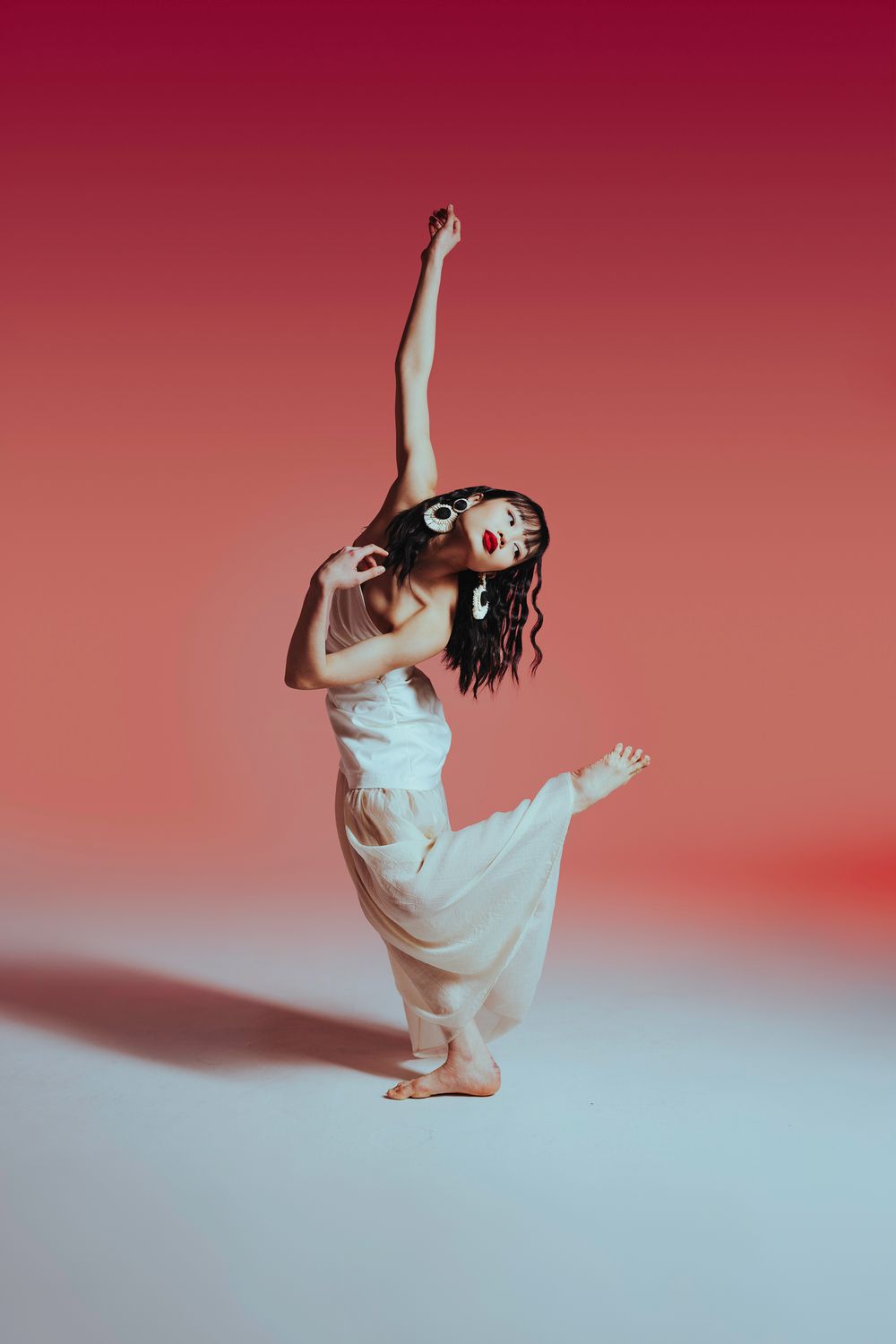Contemporary dancer in white dress performing expressive movements against red gradient background.