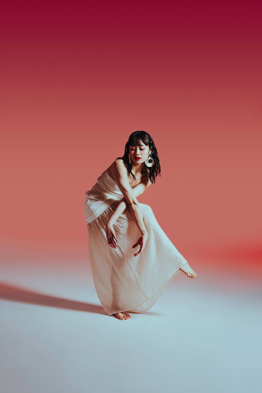 Woman in white dress floating ethereally against red and cream gradient backdrop in artistic photo series.