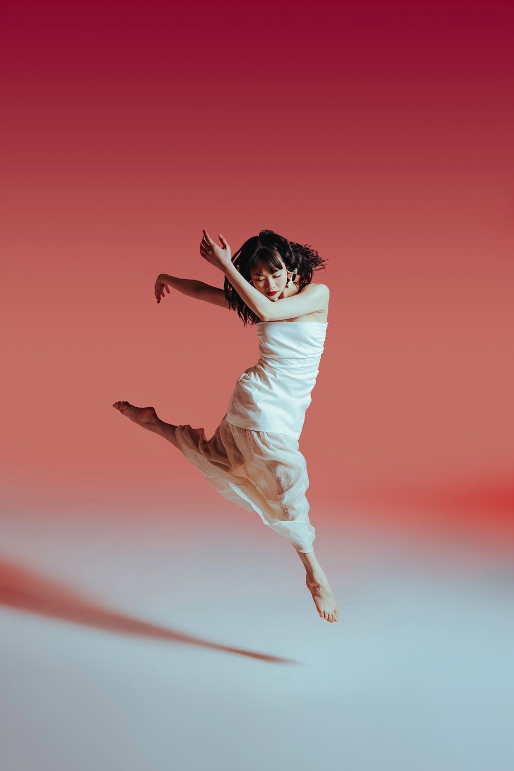 Dancer in flowing white dress leaps gracefully through the air against a dramatic red and white gradient background.