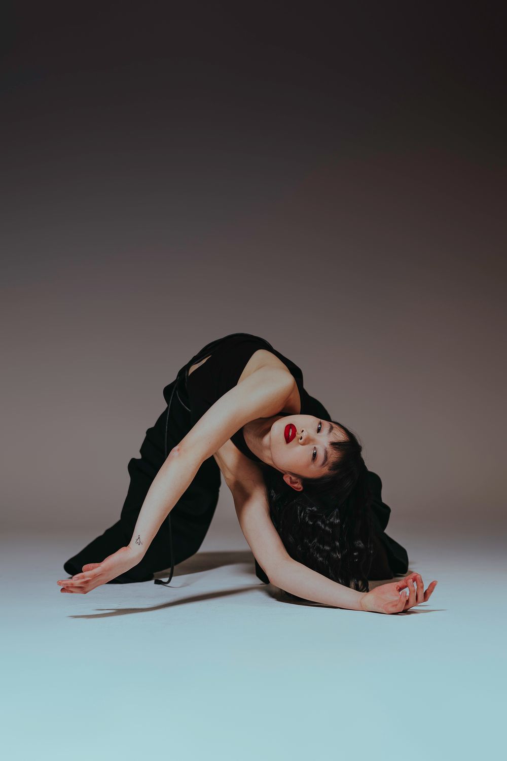 Dancer in black dress performs dramatic curved pose against gradient background in modern dance sequence.