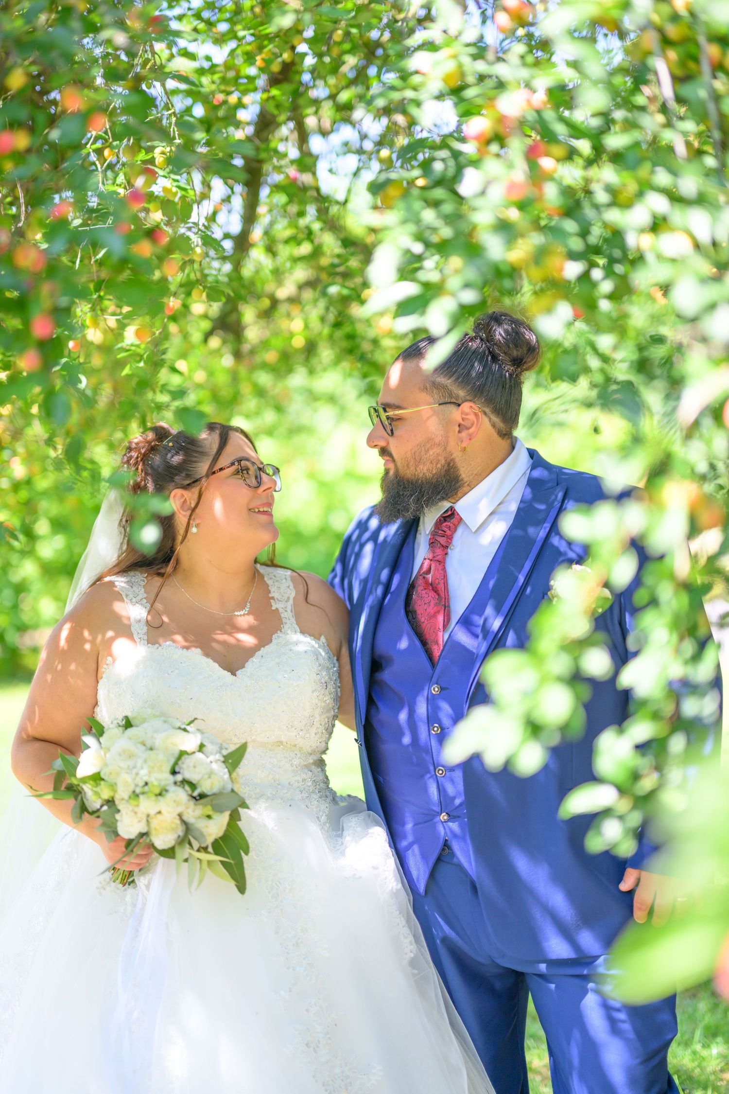 Wedding couple embraces under green apple tree branches in natural sunlight.