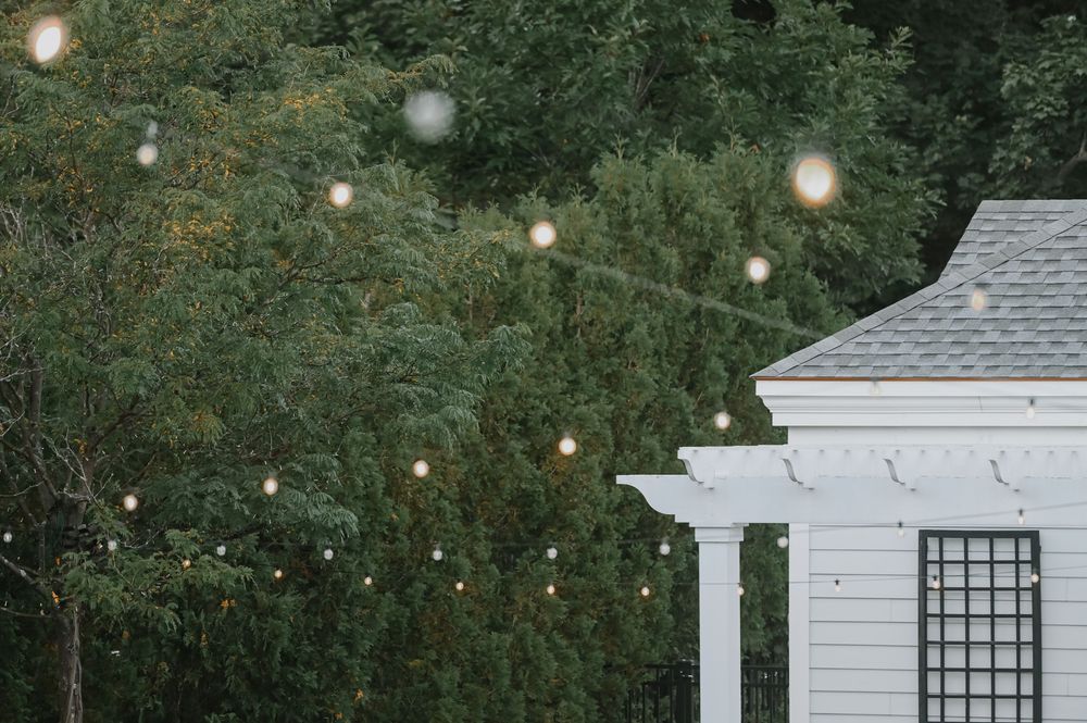 String lights illuminate a garden scene with a white house corner and trellis against lush greenery at dusk.