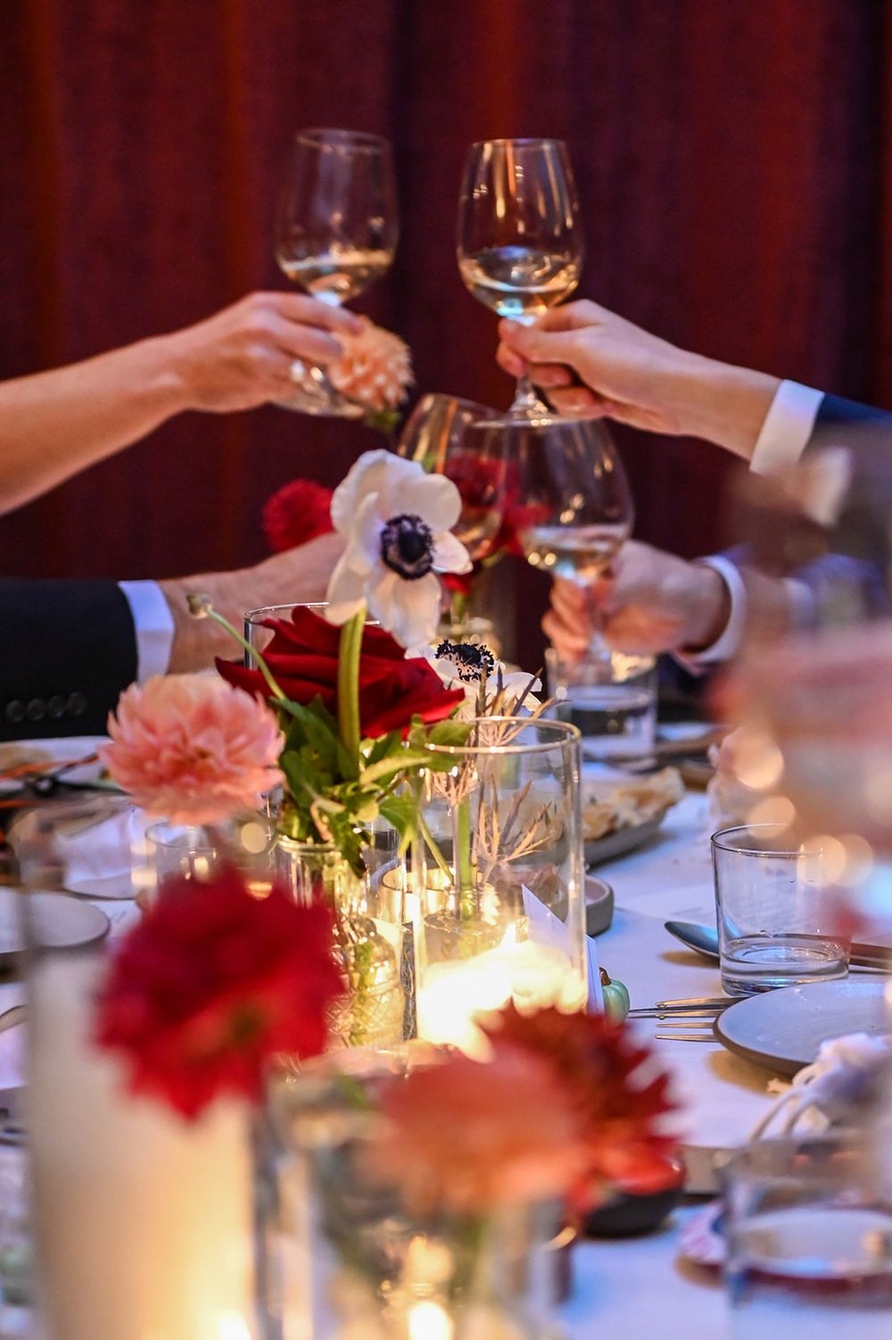 Hands raise wine glasses in a toast over an elegant table setting with red flowers and candlelight at a formal dinner party.