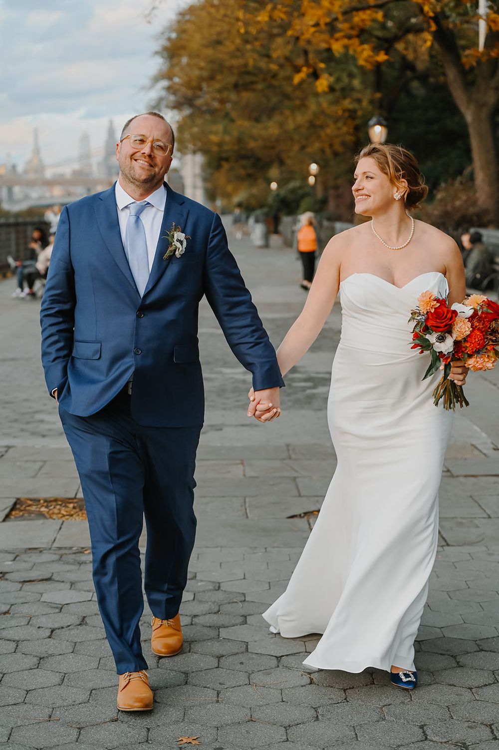 A bride in white gown and groom in navy suit walk hand in hand along a stone path lined with autumn trees.
