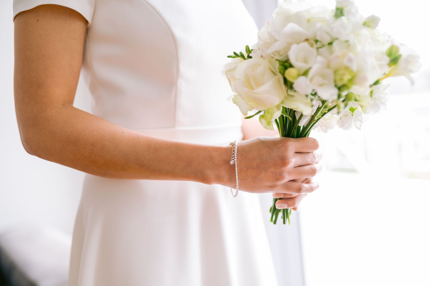 Elegant bride in white dress holding a delicate bouquet of white flowers and greenery against bright background.