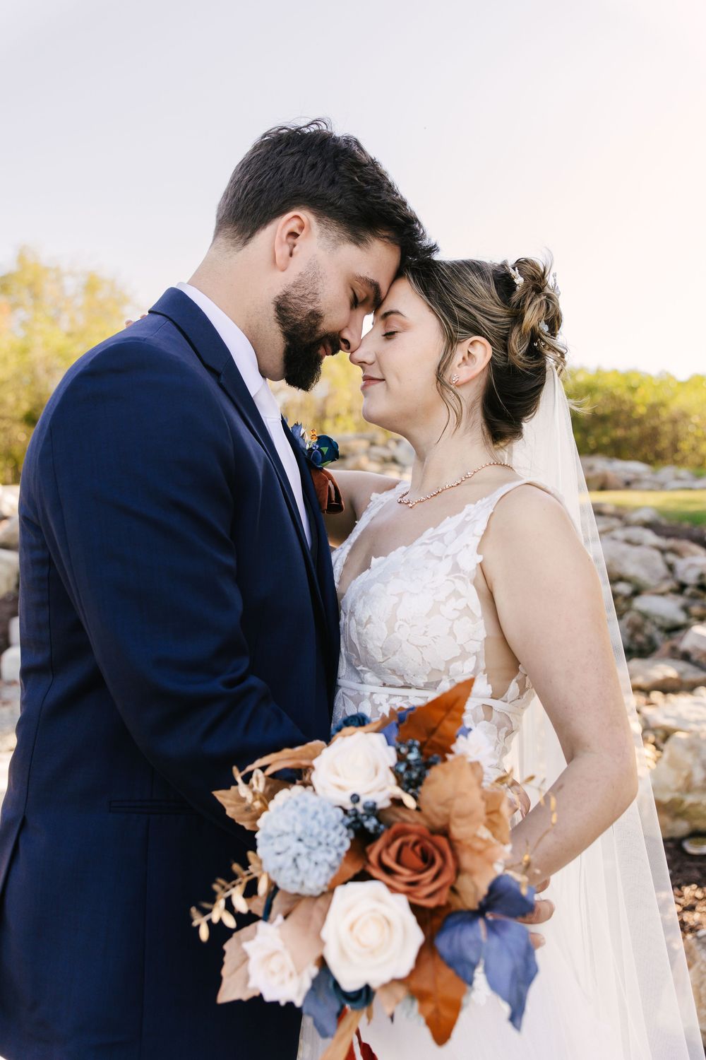 Romantic outdoor wedding photos showing couple embracing with autumn bouquet of peach and blue flowers at sunset.