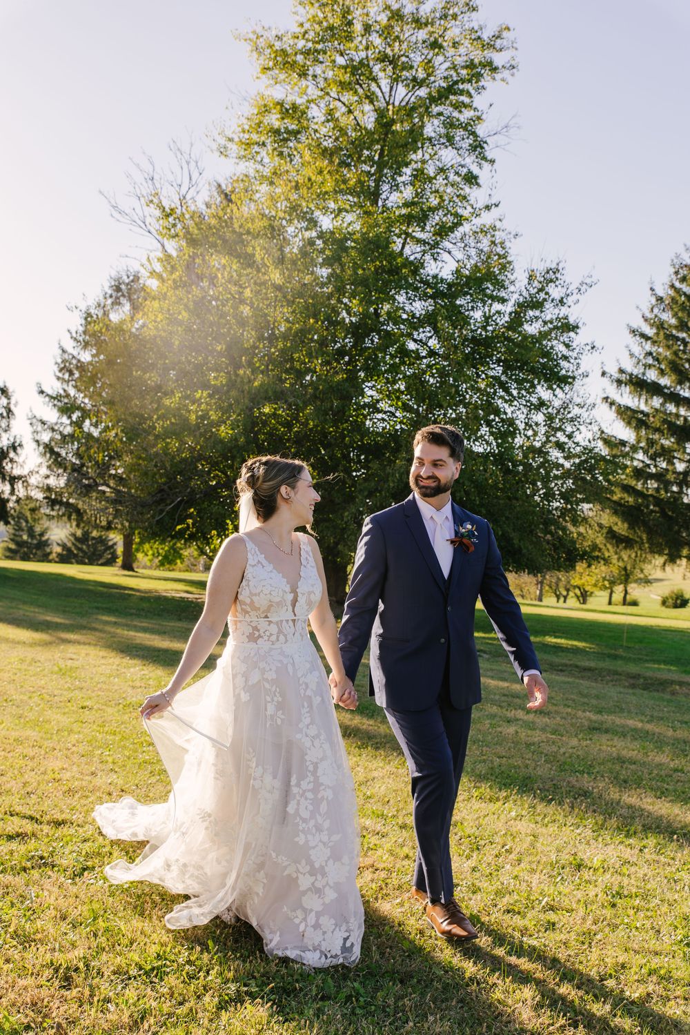 A couple in wedding attire walks together on a sunlit grassy hill during golden hour.