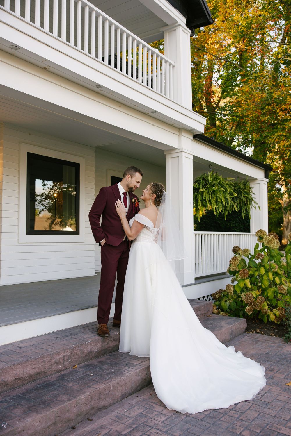 A romantic first look moment unfolds on a white columned porch with autumn foliage in the background.