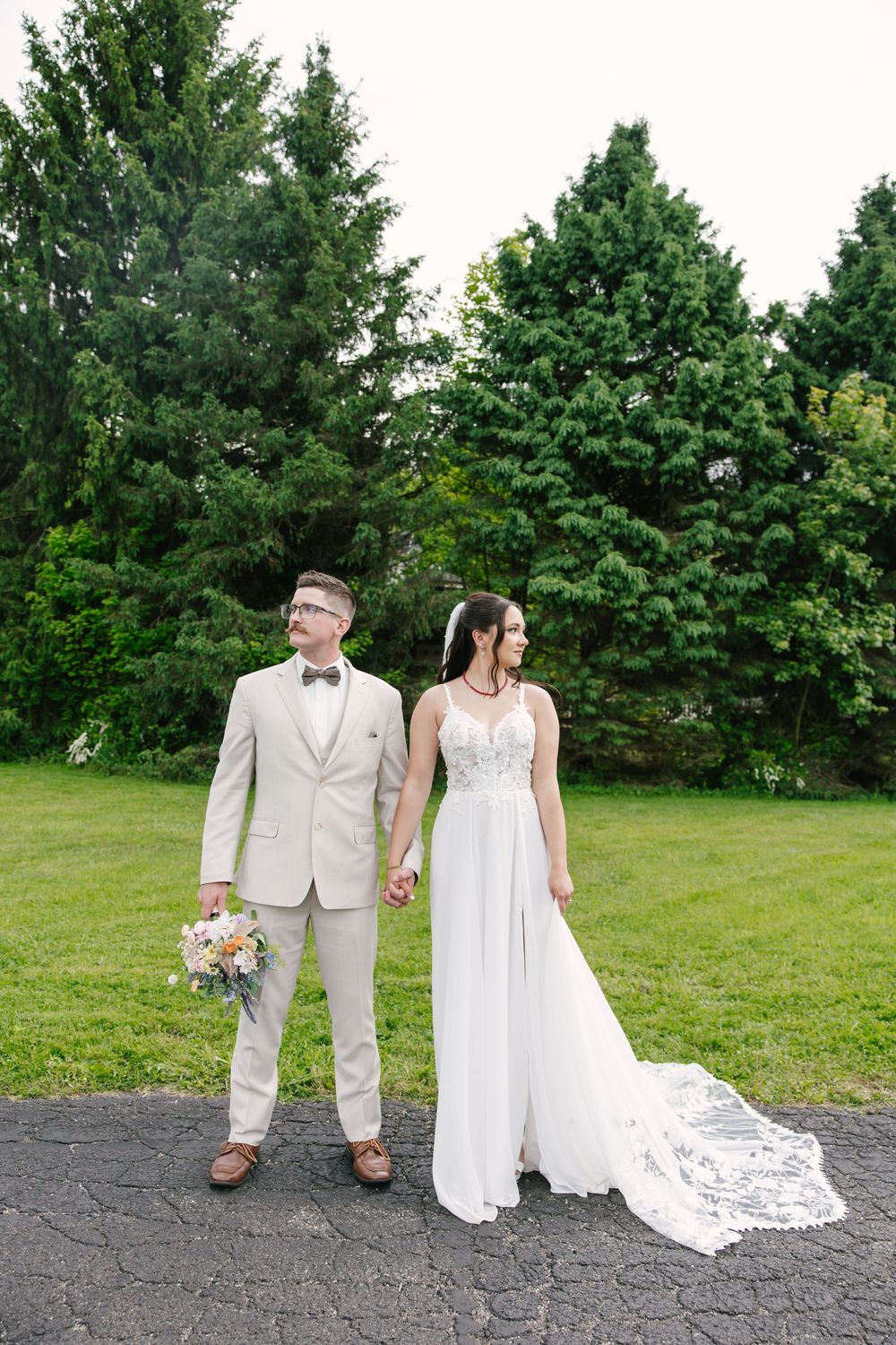 A bride and groom hold hands outdoors in a lush green setting, she in a white gown, he in a beige suit with bow tie.