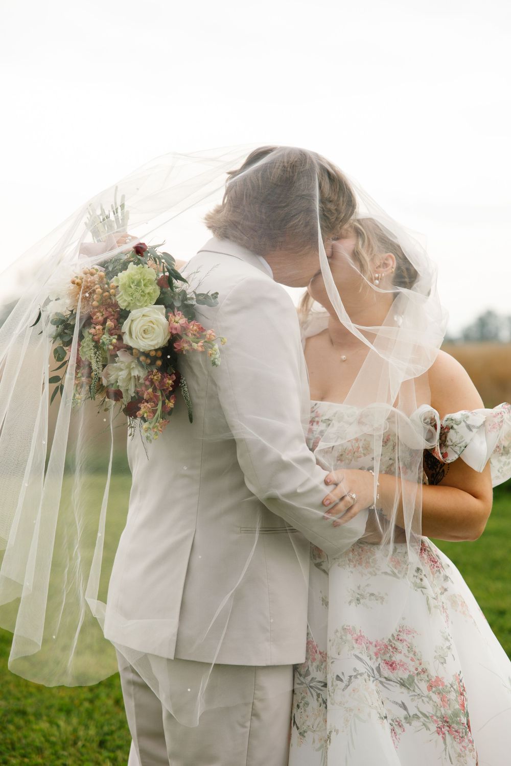 Bride and groom kissing under a flowing veil outdoors, bride holding a floral bouquet in a floral wedding dress.