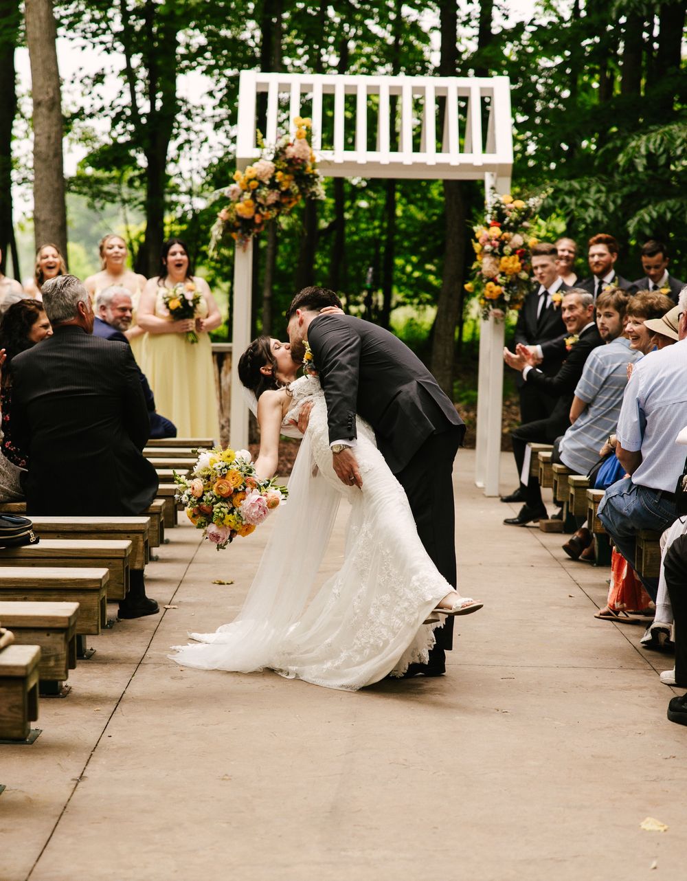 Outdoor wedding ceremony with white gazebo and wooden benches surrounded by trees.