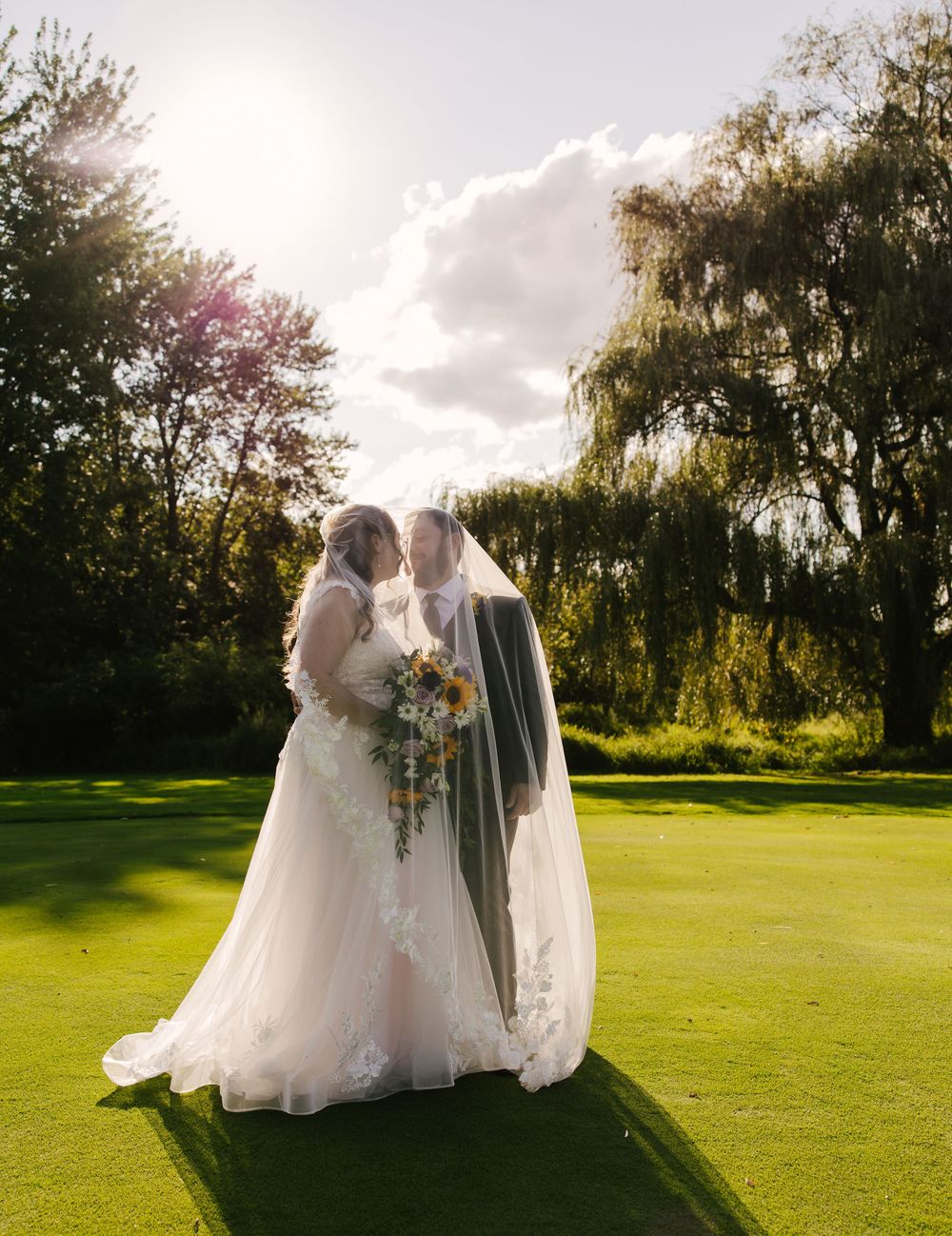 Wedding couple poses on sunny golf course with flowing white dress and bouquet.