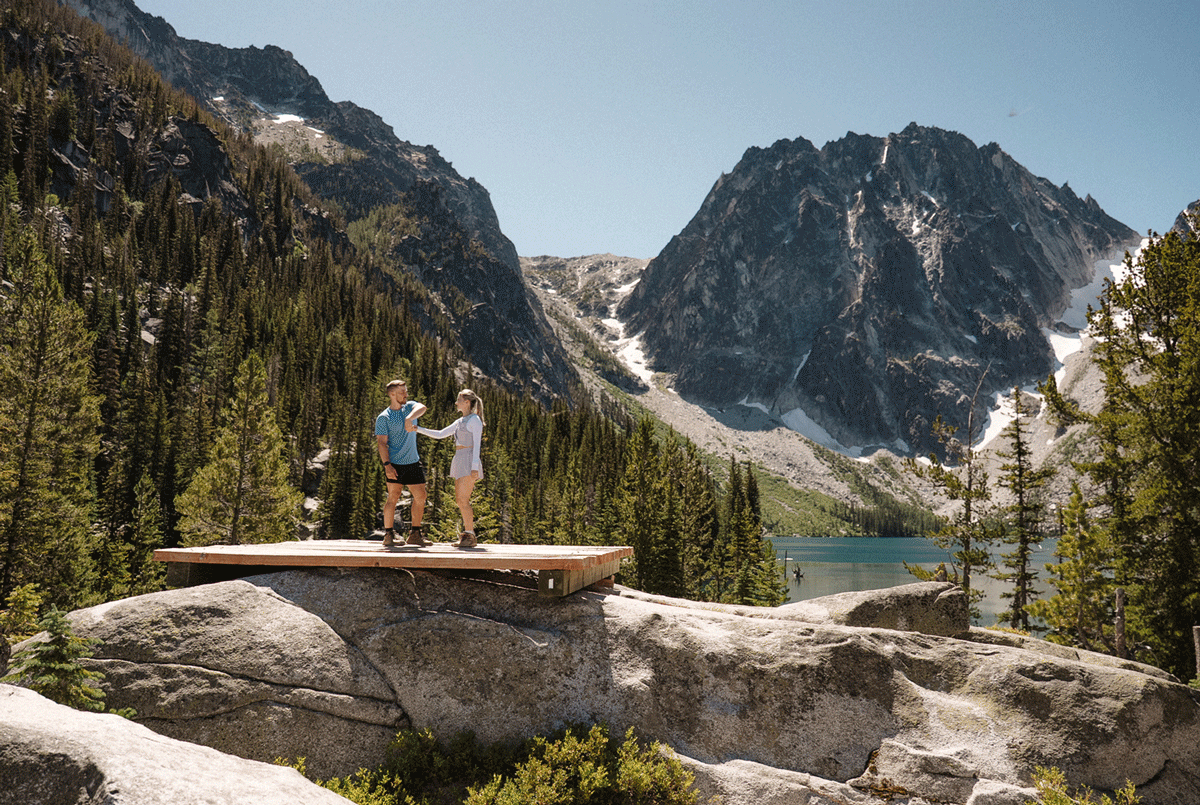 Two people stand on a rocky outcrop overlooking Colchuck Lake and mountain peaks in the Cascade Range.
