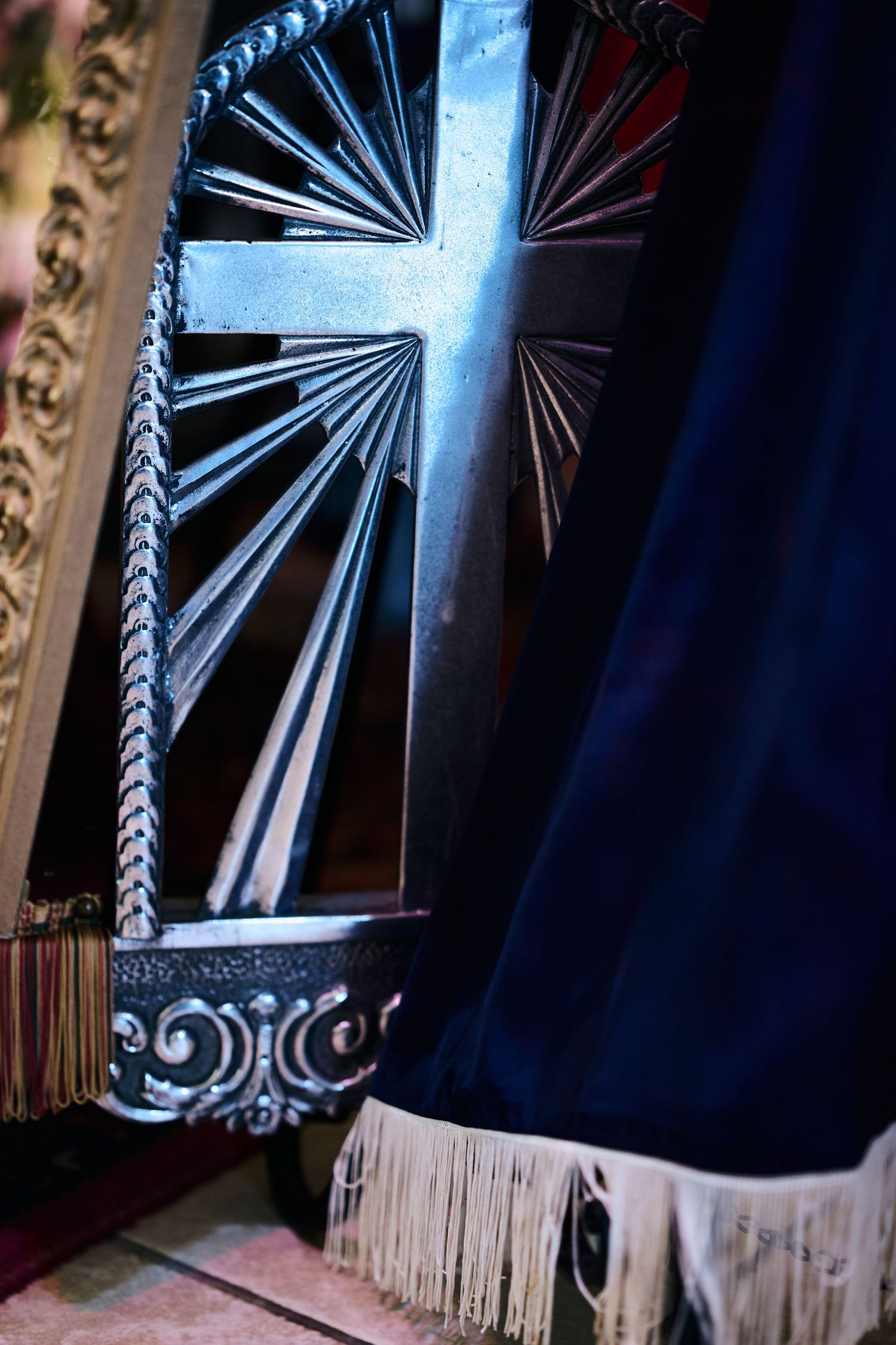 An ornate silver cross with radiating lines is displayed against a dark blue curtain with fringe.
