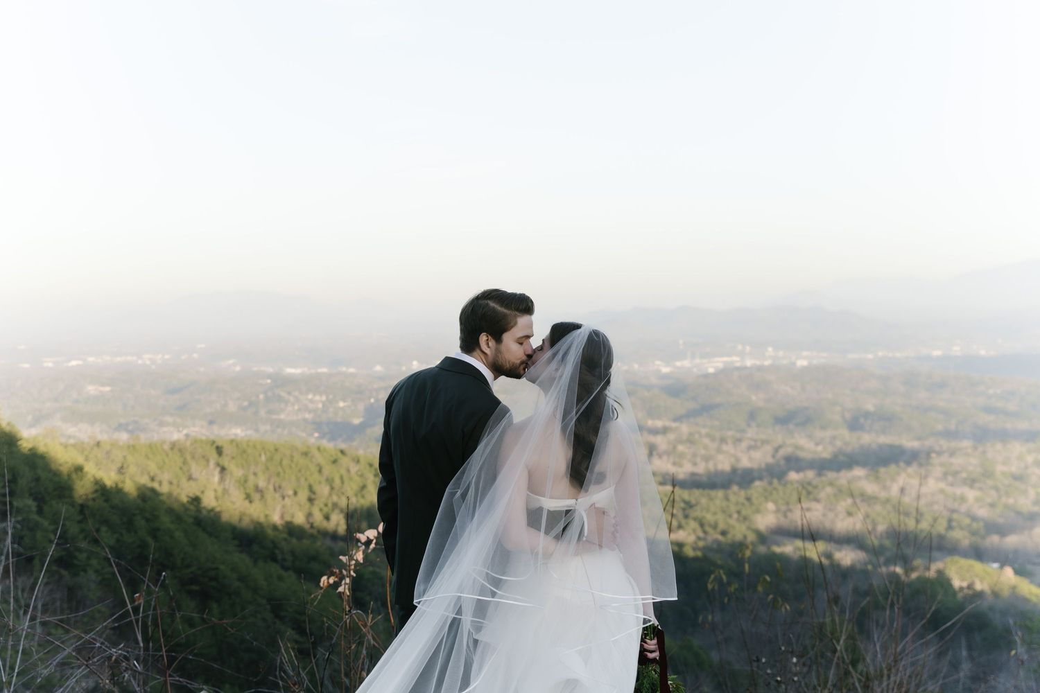 A couple dances together on a lush green hilltop surrounded by trees on a sunny summer day.