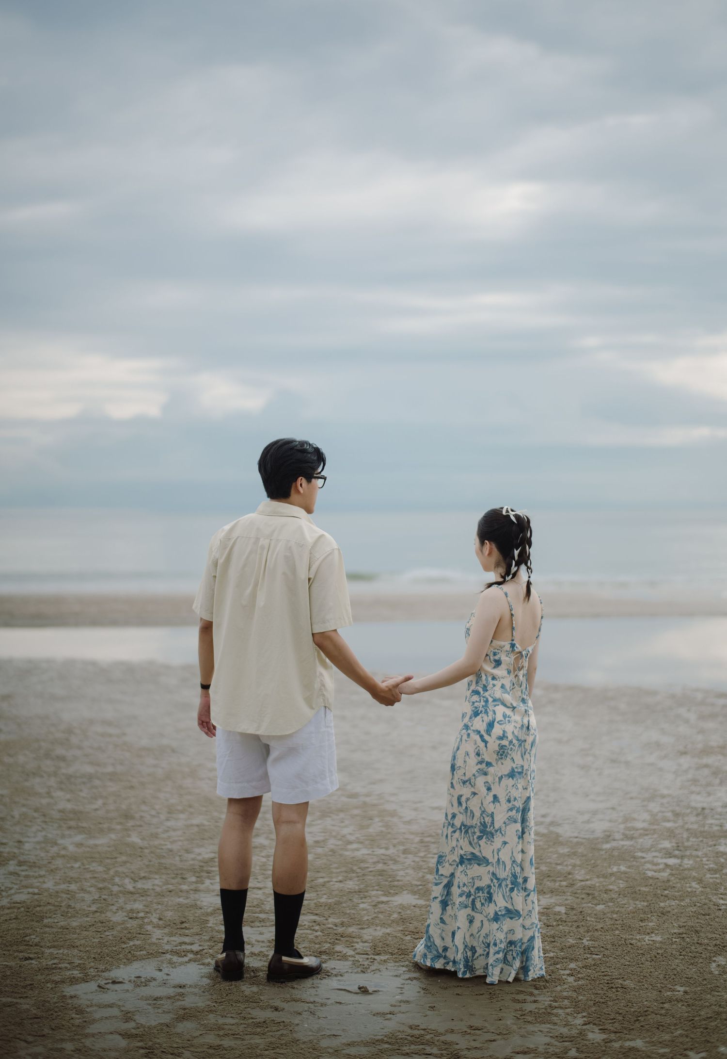 A couple walks hand in hand on a misty beach wearing vintage-inspired clothing.