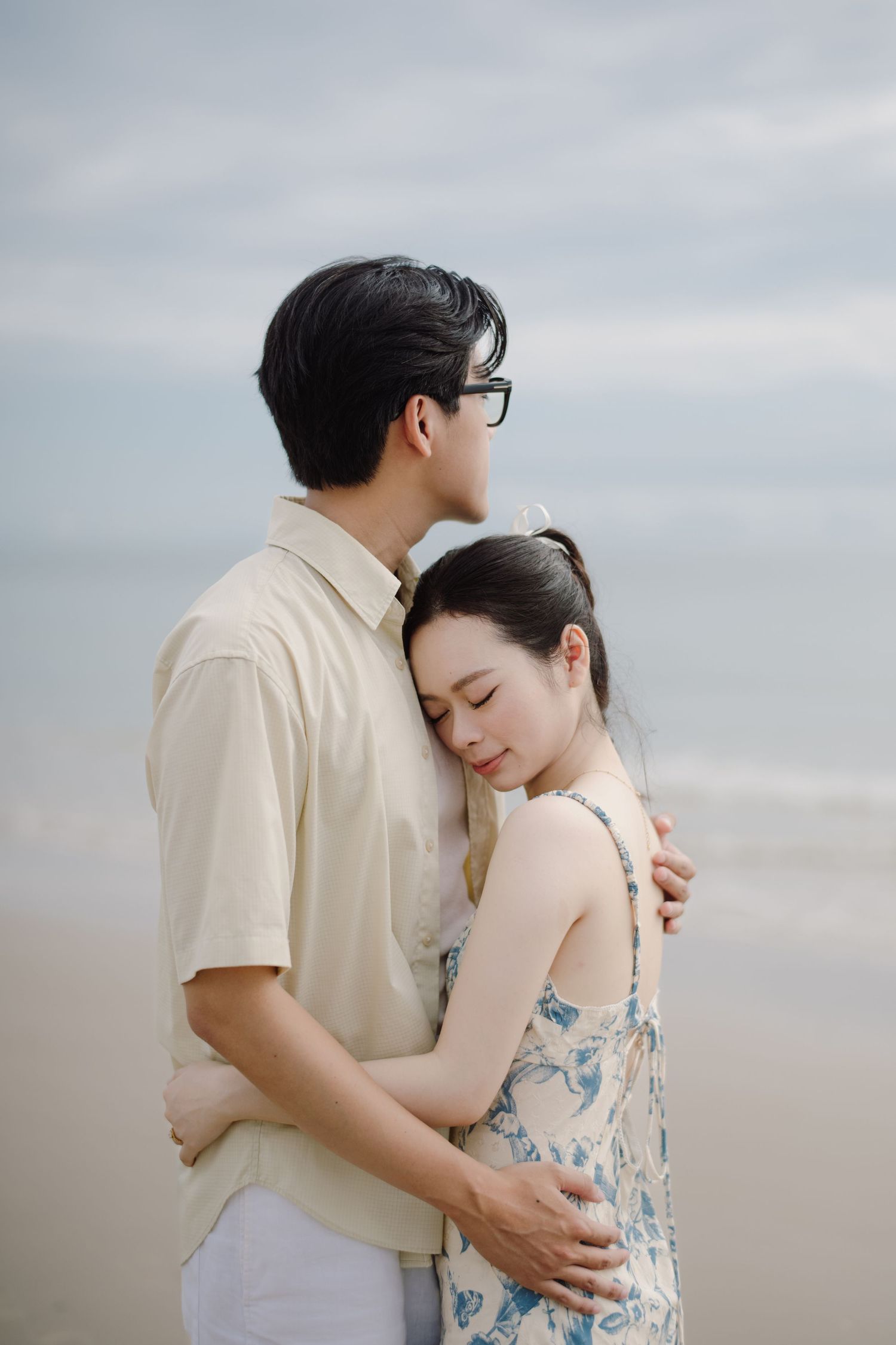 Couple in white and floral dress share intimate moment against overcast sky.