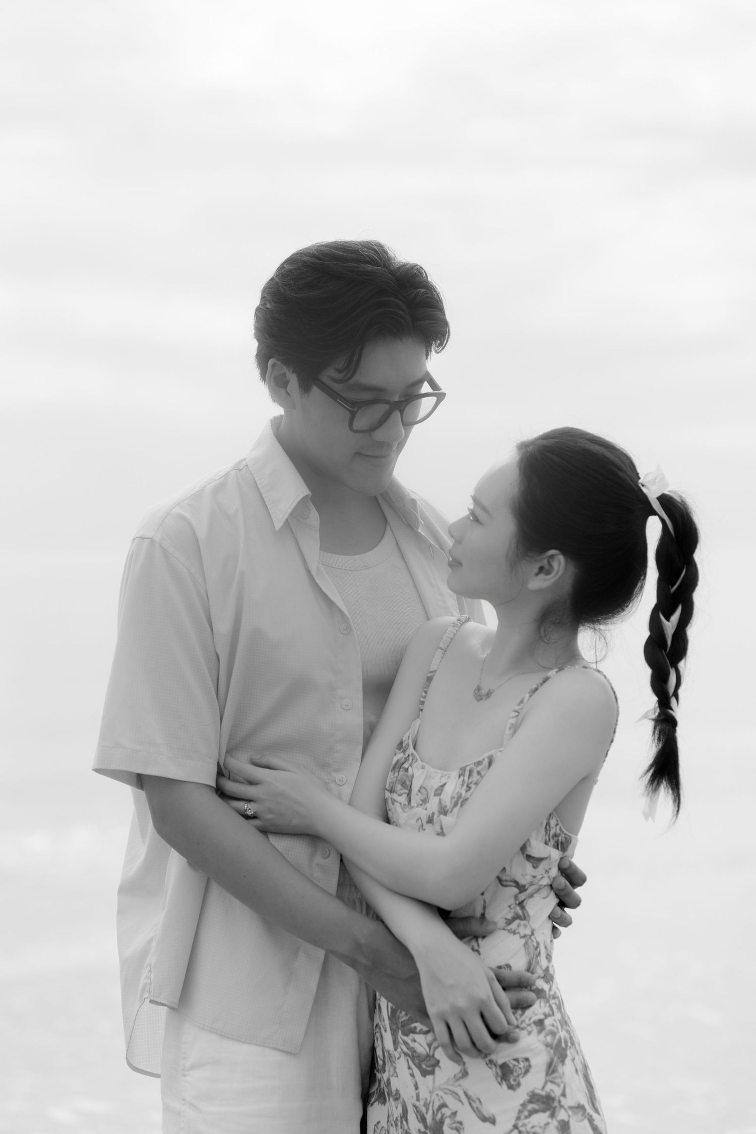 Black and white romantic beach photoshoot showing tender embrace against cloudy sky.
