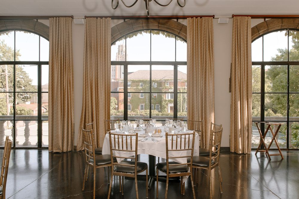 A panoramic view of an elegant dining room with large arched windows, cream curtains, and round tables set for service.