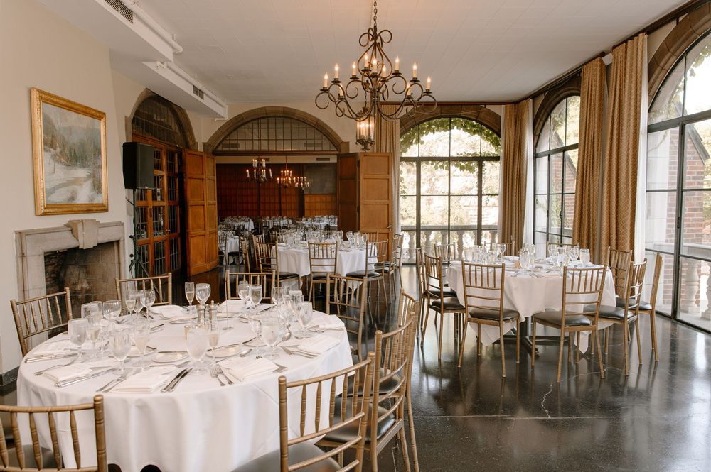 Elegant dining room with round tables, white linens, gold chiavari chairs, and arched windows in a classic event venue.