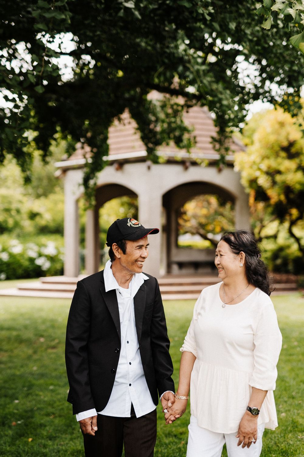 A couple wearing black and white outfits hold hands and smile at each other while walking in a park with arches.