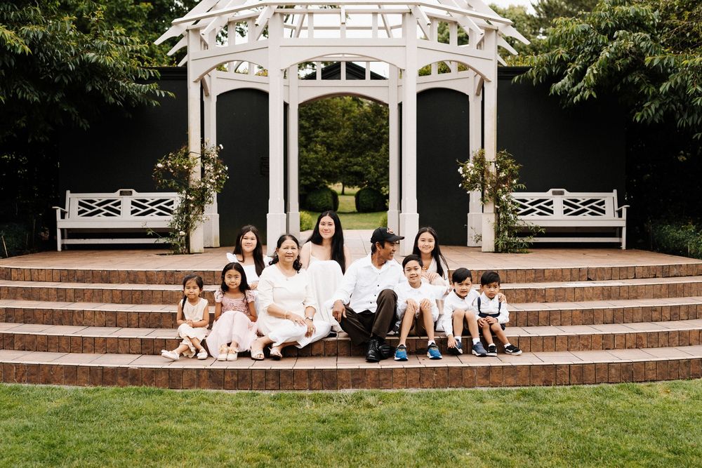 Group in white clothing sitting on concrete stairs outside ornate gazebo for outdoor portrait session.