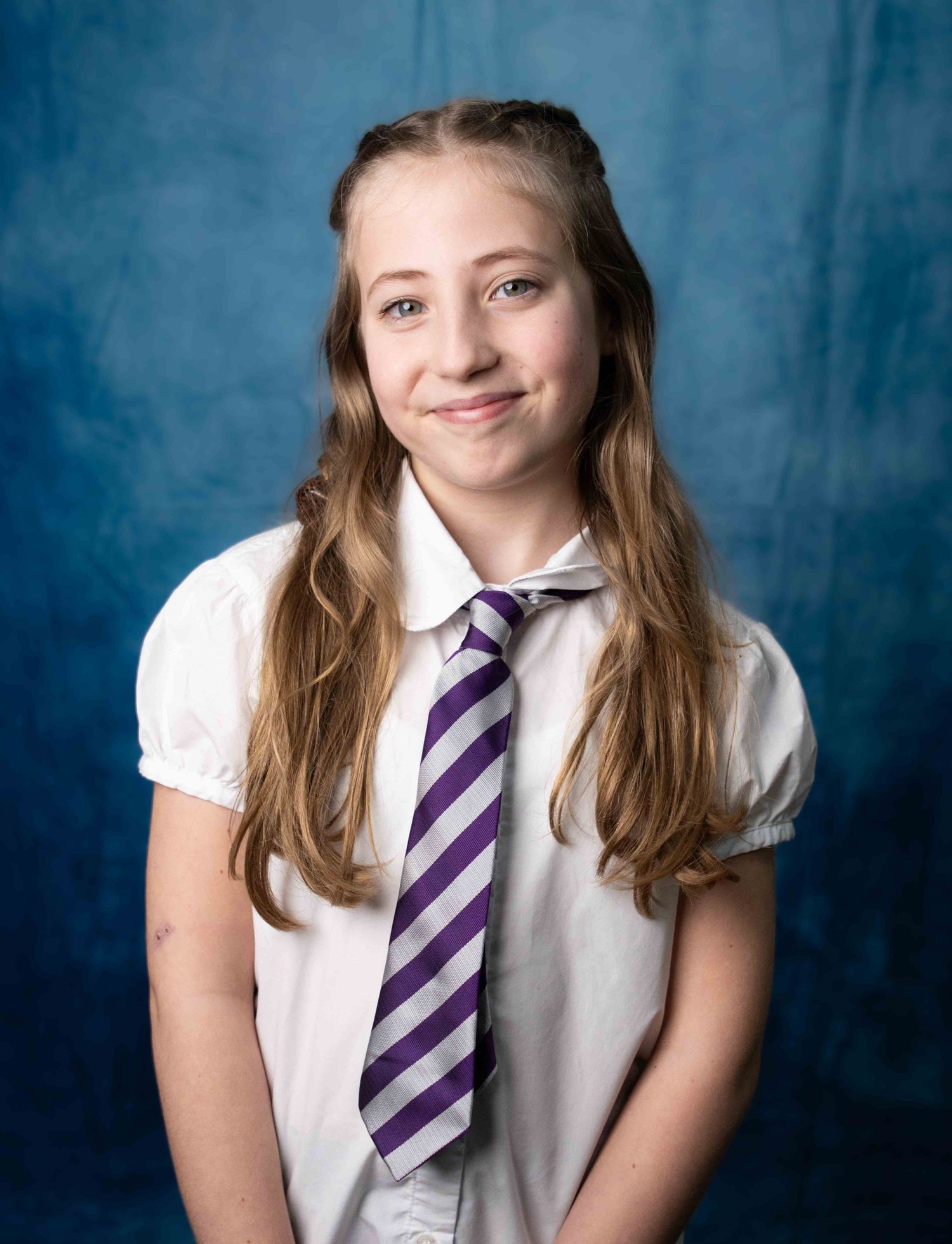 Student portrait against blue background wearing white blouse and purple striped tie in school uniform style.