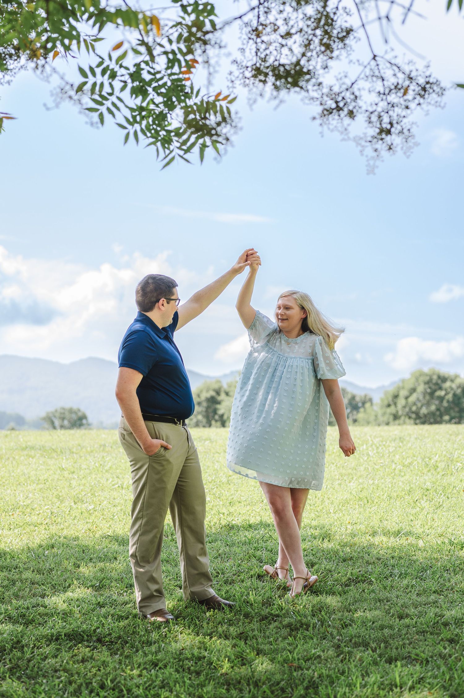 A couple stands together in a sunny field against mountain backdrop as they hold hands and dance on the grass.