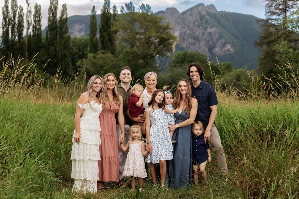 Large family group portrait smiling outdoors in a lush green meadow with mountain backdrop on a cloudy day.