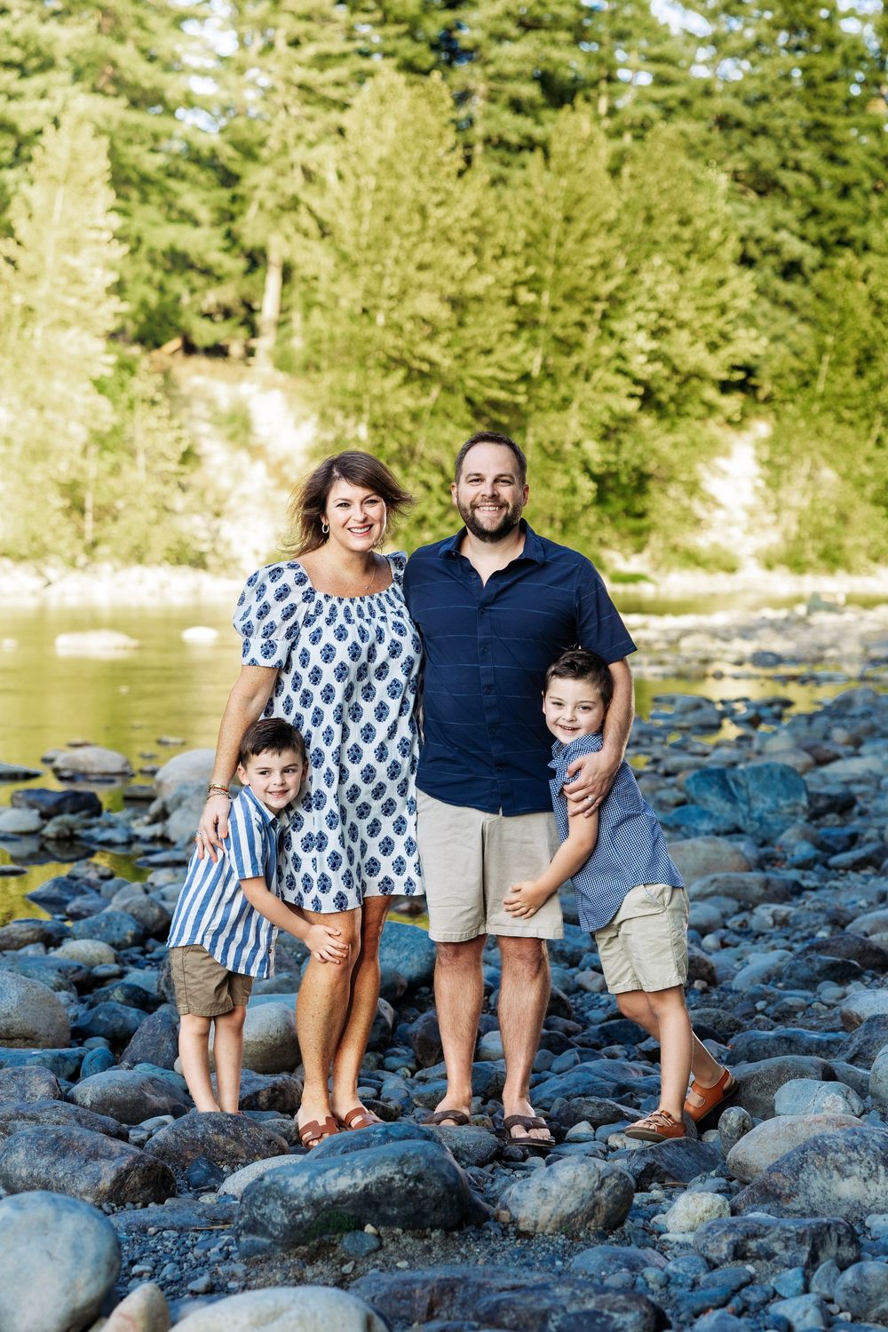 Family of four posing on rocky riverbank surrounded by lush green trees during a sunny outdoor photo session.