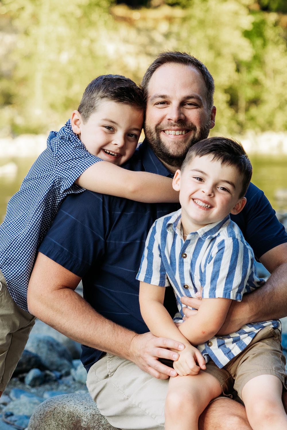 A smiling father sits outdoors with his two young sons hugging him, near a river with green trees in background.