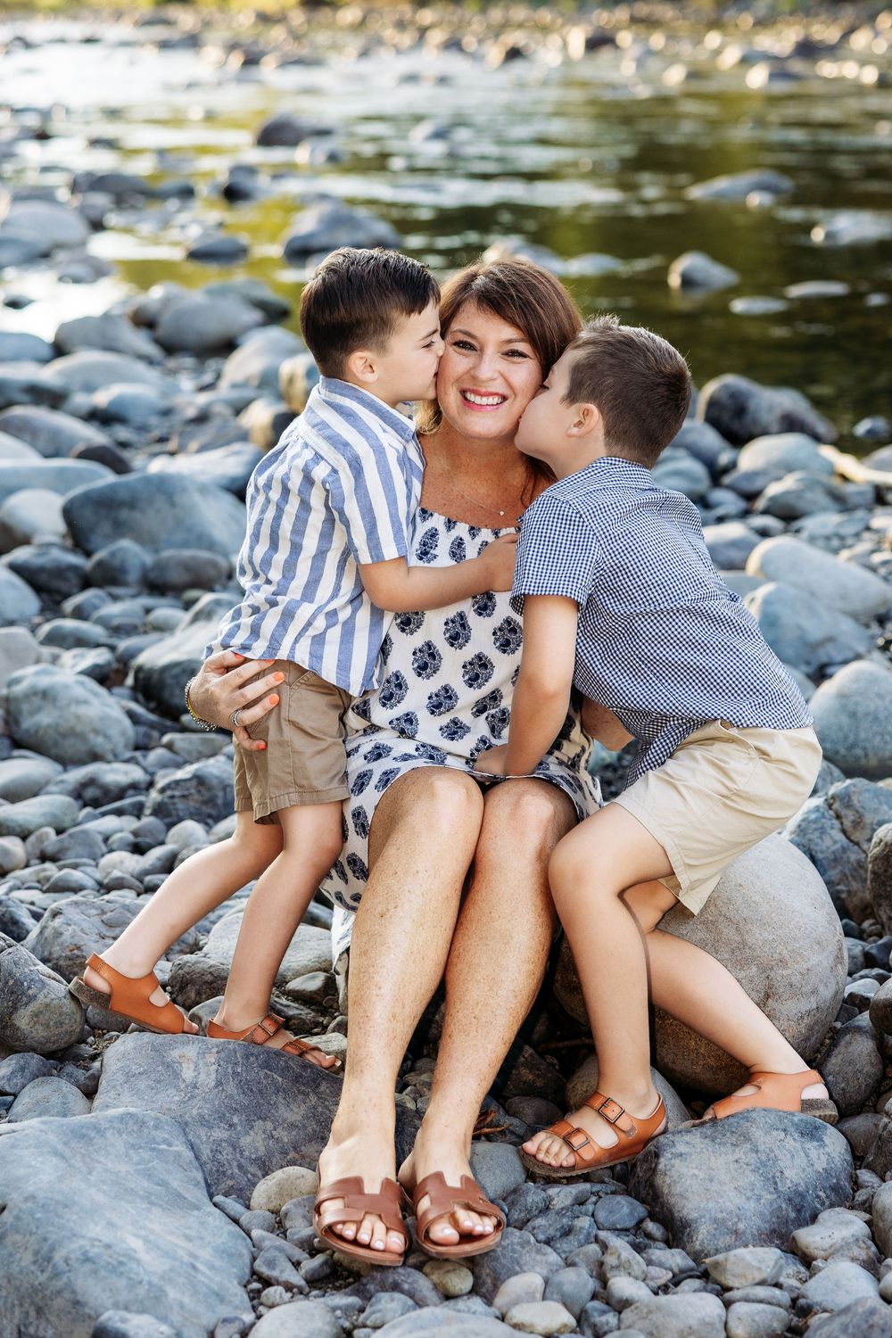 A mother sits on rocks by a river, smiling as her two young sons in striped shirts kiss her cheeks.