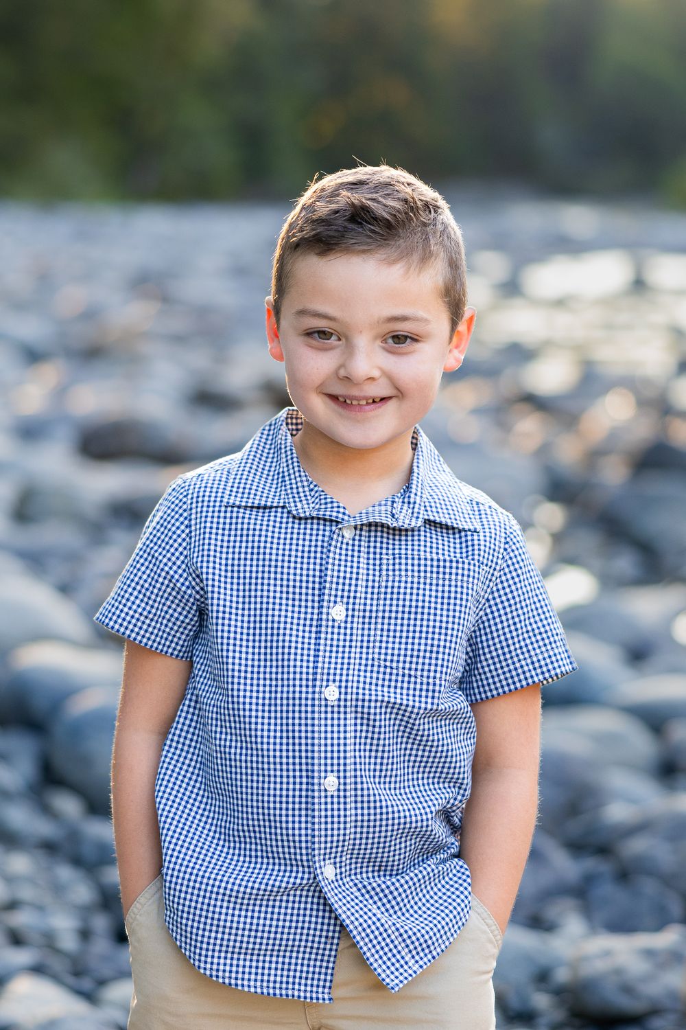 A smiling young boy in a blue gingham shirt stands confidently on a rocky outdoor background.