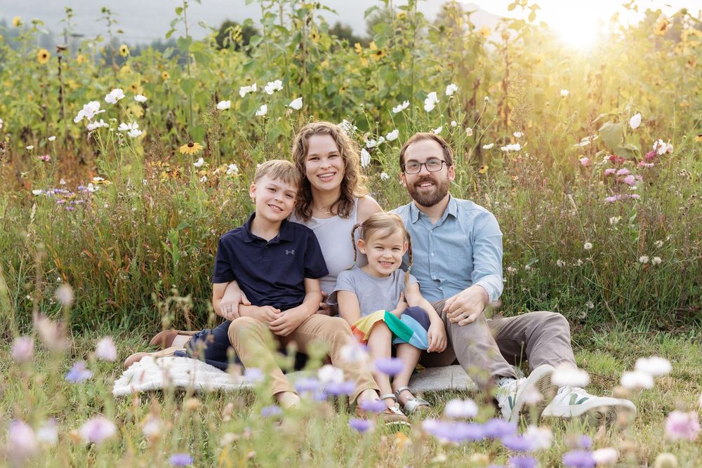 Family of four smiling together in a sunny wildflower meadow, parents and two children enjoying an outdoor portrait session.