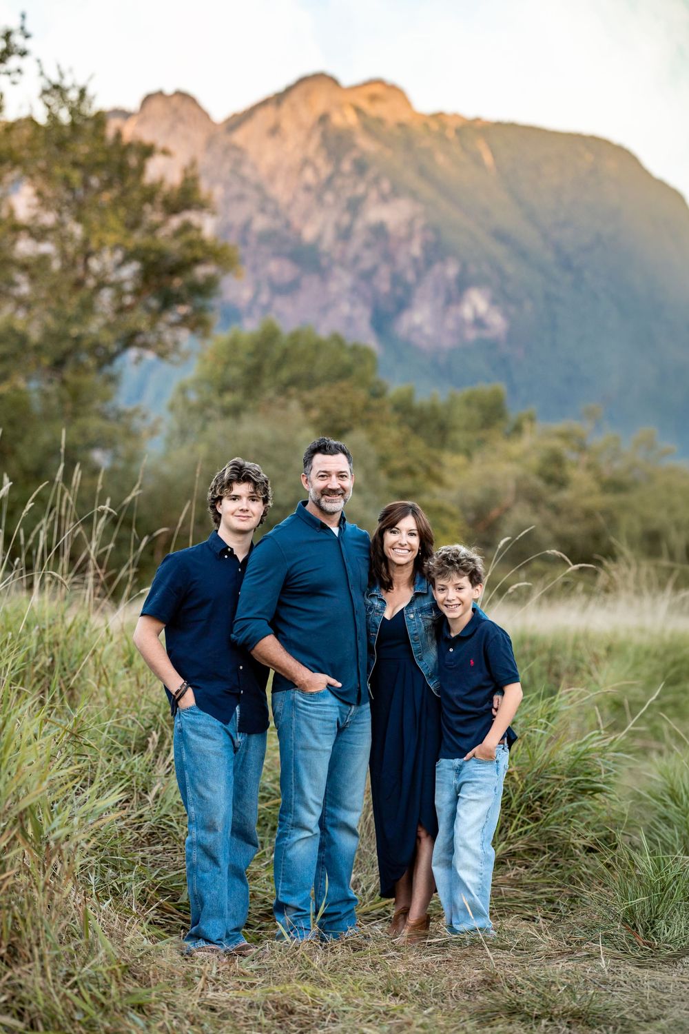 Family of four posing outdoors in navy blue outfits with a scenic mountain and green field backdrop.