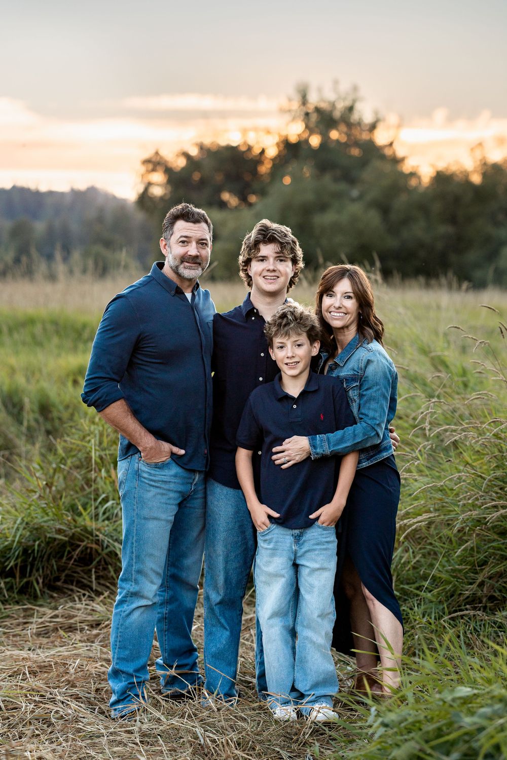 Family of four poses in a golden field at sunset, wearing coordinated navy and denim outfits.