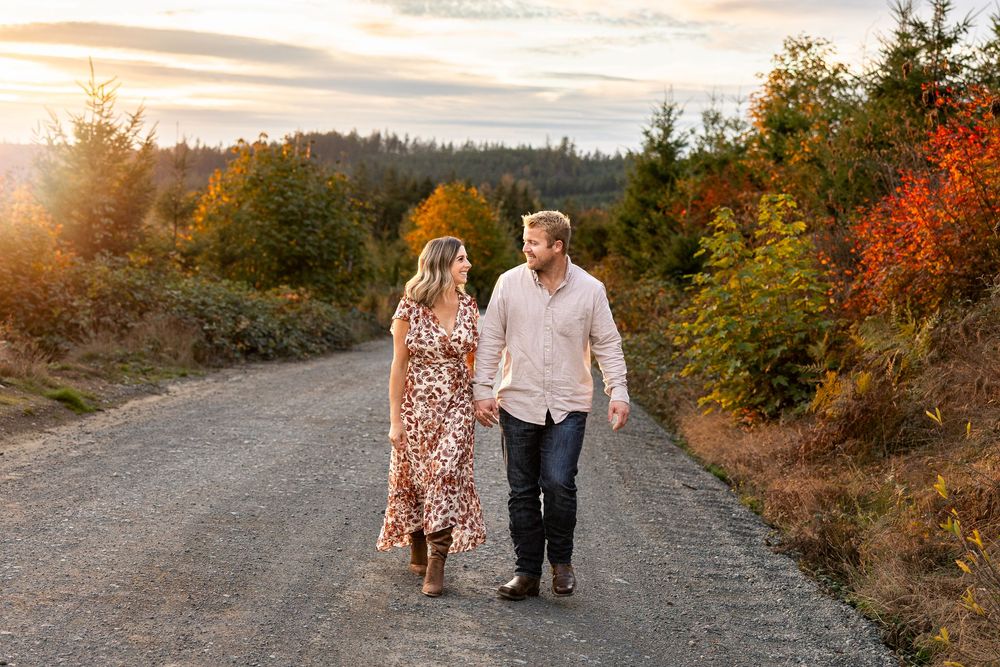 Couple holding hands walking on a gravel path surrounded by colorful autumn foliage at golden hour.