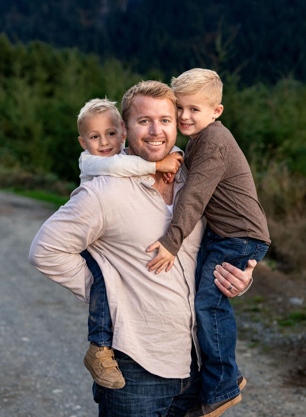 A smiling man carries two young blonde boys on his back and front outdoors on a scenic tree-lined gravel path.