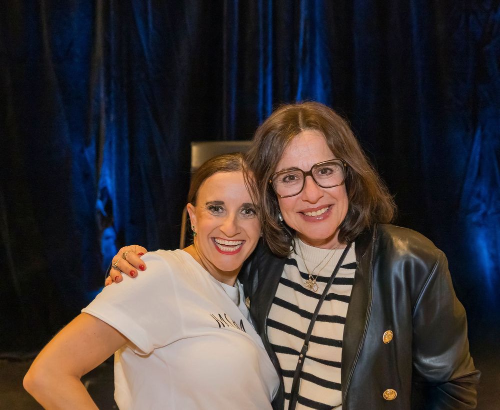 Two people pose together smiling at a professional event with blue curtains in the background.