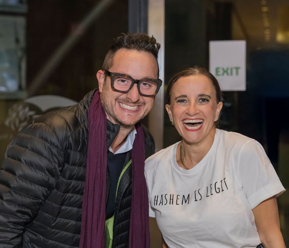 Two people smiling together at a casual event, one wearing a puffy jacket and scarf, the other in a white t-shirt.