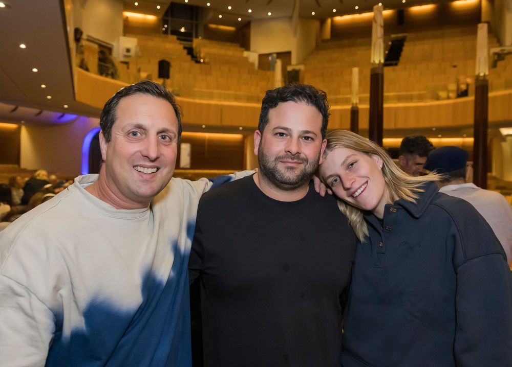Three people pose together smiling at a performance venue with warm lighting and theater seating visible in background.