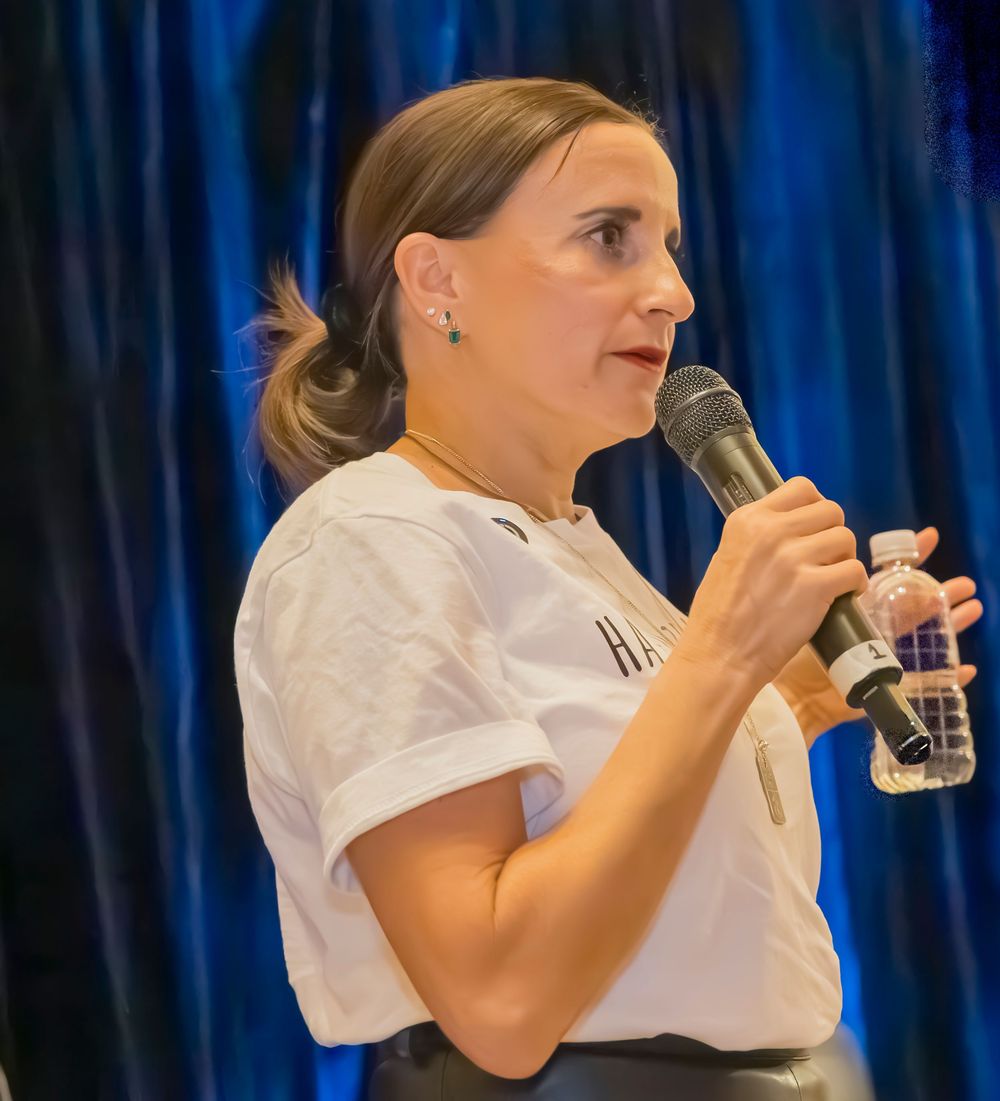 Female performer in white shirt holds microphone while singing on stage against blue curtain backdrop.