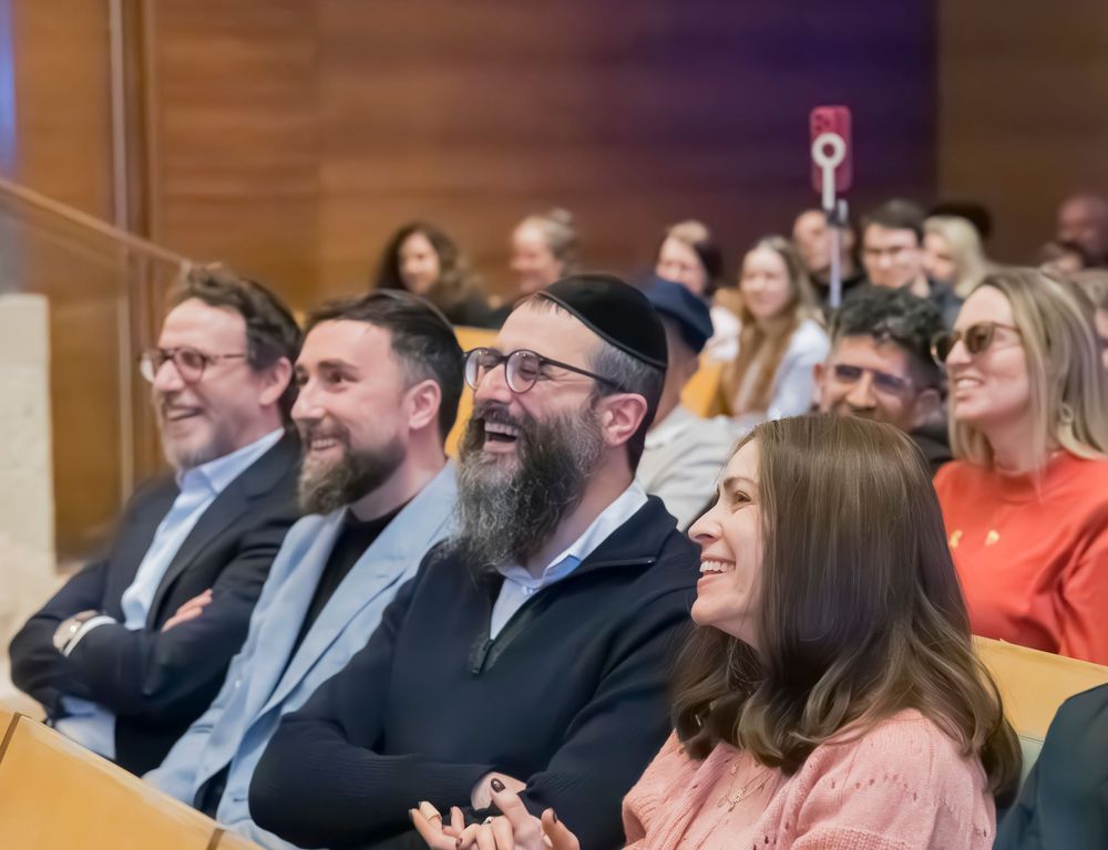 Conference attendees sit in an auditorium reacting with smiles and amusement during a presentation.