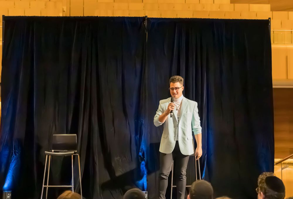 A performer in a light colored blazer stands on stage with a microphone against a dark curtain backdrop.