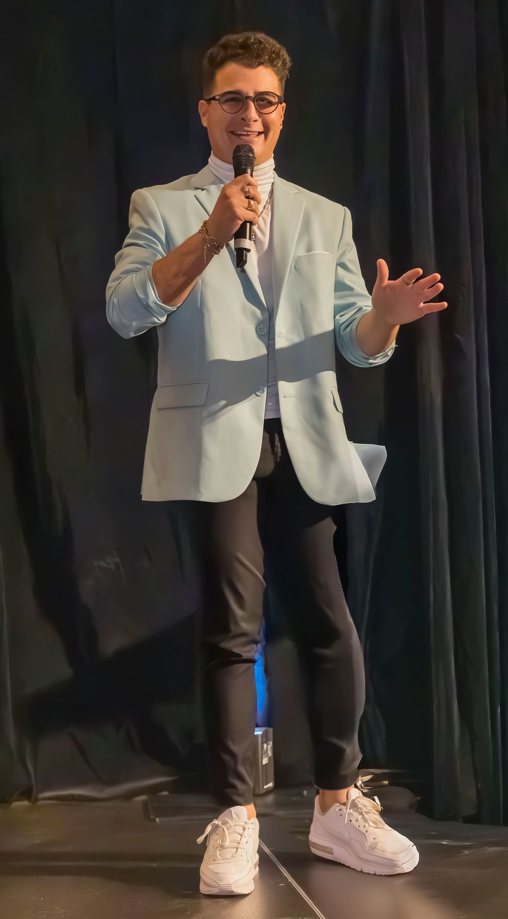 A performer in a light blue blazer, dark pants and white sneakers speaks into a microphone on stage.