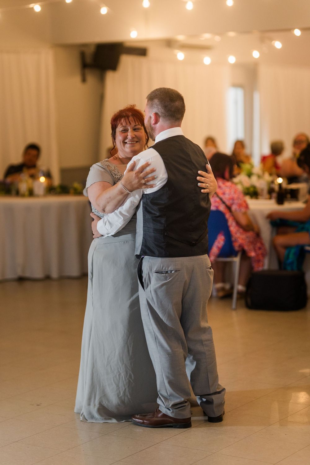 A tender dance moment unfolds on a reception hall floor illuminated by warm string lights above.
