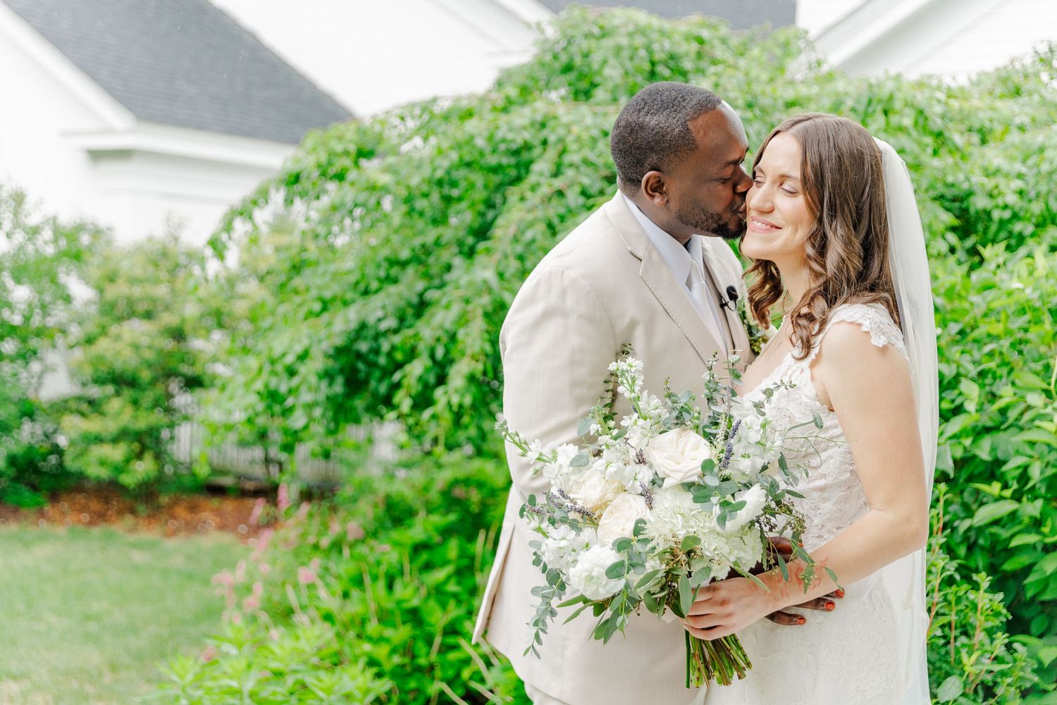 A joyful wedding couple shares candid laughter while posing together on a bright white porch surrounded by greenery.