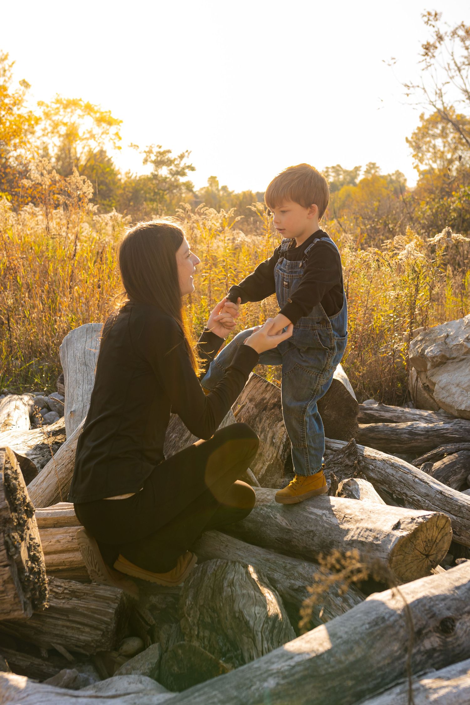 Tender moment between mother and son on pile of fallen logs at sunset with golden autumn leaves surrounding them.